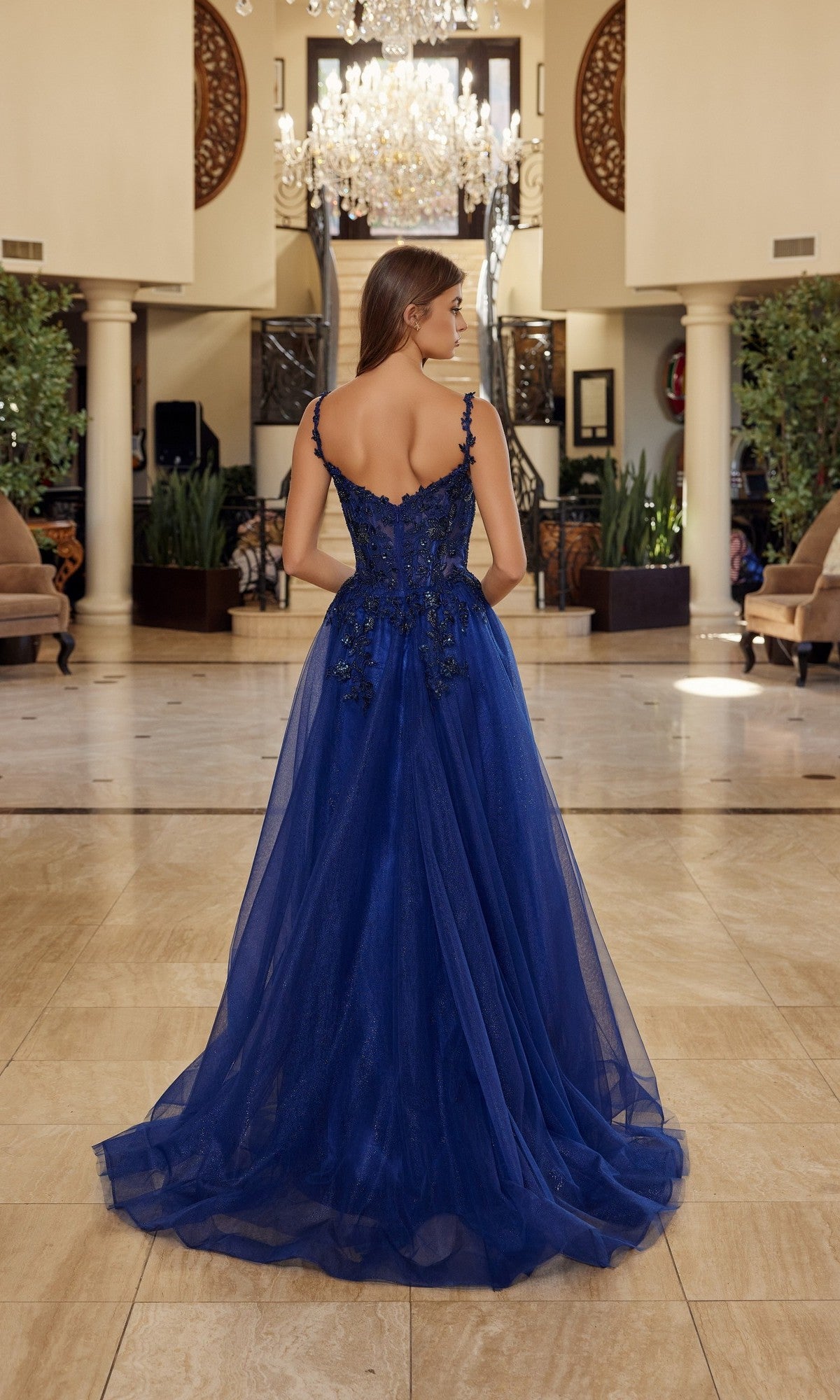 A woman wearing the Nox Anabel R1824 long prom dress, featuring a blue floral embroidered corset and sleeveless floor-length design, stands indoors beneath a chandelier in an elegant, spacious room, facing away.